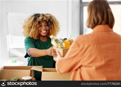 charity, donation and volunteering concept - happy smiling female volunteer and woman taking box of food at distribution or refugee assistance center. volunteer giving food at refugee assistance center