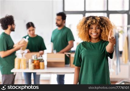 charity, donation and volunteering concept - happy smiling female volunteer pointing to camera and international group of people packing food in boxes at distribution or refugee assistance center. happy volunteers packing food in donation boxes