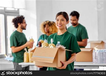 charity, donation and volunteering concept - happy smiling female volunteer with food in box and international group of people at distribution or refugee assistance center. happy volunteers packing food in donation boxes
