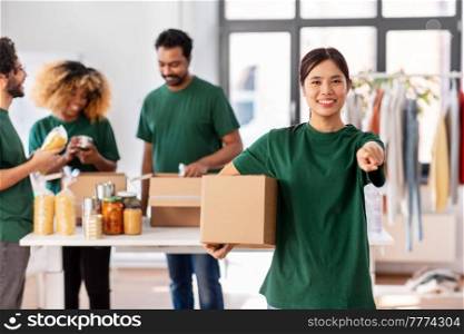 charity, donation and volunteering concept - happy smiling female volunteer with food in box pointing to camera over international group of people at distribution or refugee assistance center. female volunteer with food in box points to camera
