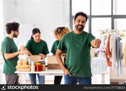 charity, donation and volunteering concept - happy smiling male volunteer pointing to camera and international group of people packing food in boxes at distribution or refugee assistance center. happy volunteers packing food in donation boxes