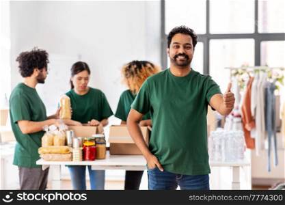 charity, donation and volunteering concept - happy smiling male volunteer showing thumbs up gesture and international group of people packing food in boxes at distribution or refugee assistance center. happy volunteers packing food in donation boxes