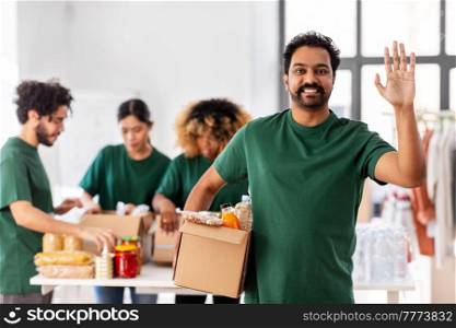 charity, donation and volunteering concept - happy smiling male volunteer with food in box waving hand over and international group of people at distribution or refugee assistance center. male volunteer with food in box waving hand