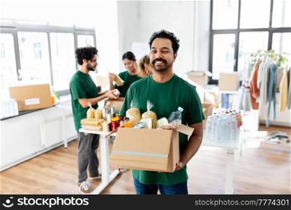charity, donation and volunteering concept - happy smiling male volunteer with food in box and international group of people at distribution or refugee assistance center. happy volunteers packing food in donation boxes
