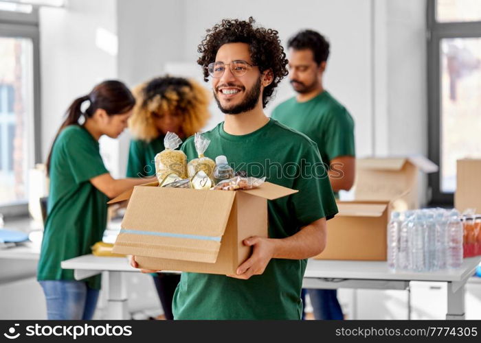 charity, donation and volunteering concept - happy smiling male volunteer with food in box and international group of people at distribution or refugee assistance center. happy volunteers packing food in donation boxes