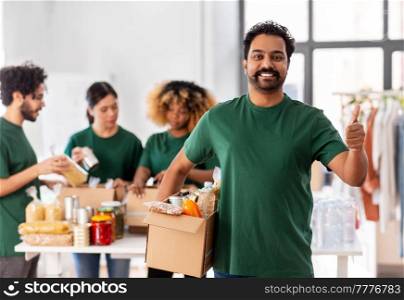 charity, donation and volunteering concept - happy smiling male volunteer with food in box showing thumbs up gesture over international group of people at distribution or refugee assistance center. male volunteer with food in box showing thumbs up