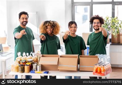 charity, donation and volunteering concept - international group of happy smiling volunteers packing food in boxes pointing to camera at distribution or refugee assistance center. volunteers packing food donation pointing to you