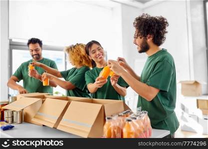 charity, donation and volunteering concept - international group of happy smiling volunteers packing food and drinks in boxes at distribution or refugee assistance center. happy volunteers packing food in donation boxes