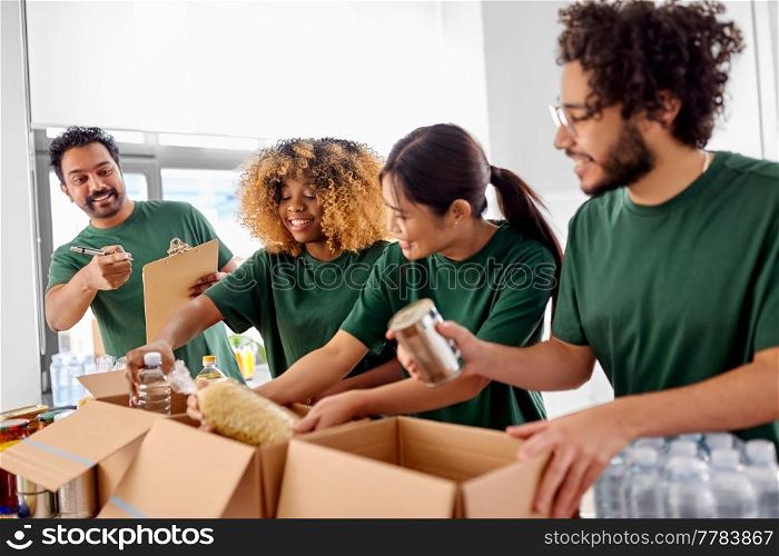charity, donation and volunteering concept - international group of happy smiling volunteers packing food in boxes according to list on clipboard at distribution or refugee assistance center. happy volunteers packing food in donation boxes