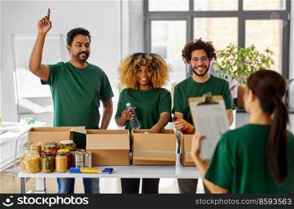 charity, donation and volunteering concept - international group of happy smiling volunteers packing food in boxes according to list on clipboard at distribution or refugee assistance center. happy volunteers packing food in donation boxes