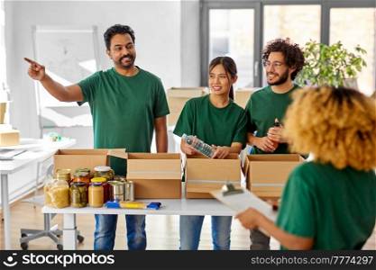 charity, donation and volunteering concept - international group of smiling volunteers packing food and drinks in boxes according to list on clipboard at distribution or refugee assistance center. happy volunteers packing food in donation boxes