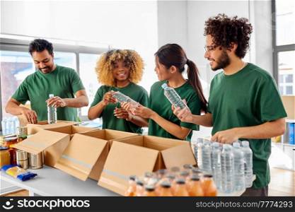 charity, food donation and volunteering concept - international group of happy smiling volunteers packing bottles of water in boxes at distribution or refugee assistance center. happy volunteers packing bottles of water in boxes