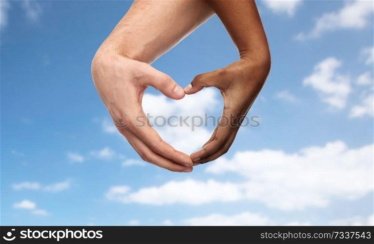 charity, love and diversity concept - close up of female and male hands of different skin color making heart shape over blue sky and clouds background. hands of different skin color making heart shape