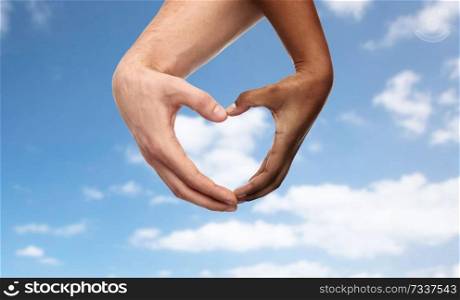 charity, love and diversity concept - close up of female and male hands of different skin color making heart shape over blue sky and clouds background. hands of different skin color making heart shape