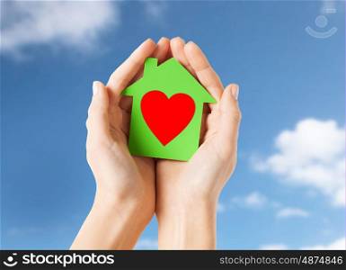 charity, real estate and love concept - close up of female hands holding green paper house with red heart shape