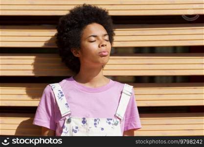 Charming African American female in casual wear standing under bright sunlight with closed eyes near wooden wall on street on summer day. Black woman standing under sunlight
