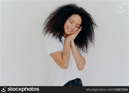 Charming beautiful woman with tender smile, leans on pressed palms, dreams about sleep, tilts head, wears white t shirt, has natural beauty, perfect teeth, poses indoor. Young dreamy Afro woman