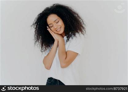 Charming beautiful woman with tender smile, leans on pressed palms, dreams about sleep, tilts head, wears white t shirt, has natural beauty, perfect teeth, poses indoor. Young dreamy Afro woman