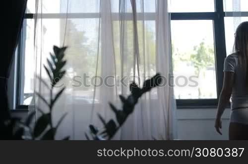 Charming blond woman with beautiful straight long hair in white underwear, t-shirt and panties, stretching near the window after early morning awakening. Back view. Stretching young sexy female exercising to get energy. Dolly shot.
