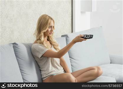 charming blonde female sitting on comfortable sofa with nude legs and panties, watching television and changing channel with remote control