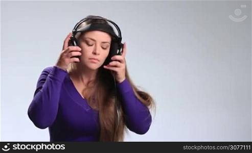 Charming brunette woman putting headphones on her ears, closing her eyes and swaying along with the music on white background. Attractive teenage girl holding earphones with both hands, opening her deep brown eyes and sheepishly smiling at the camera.