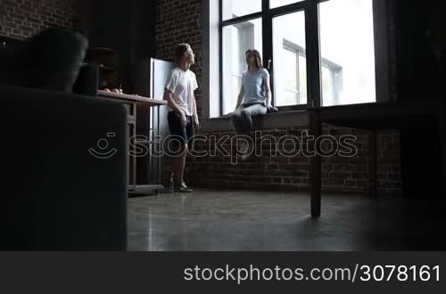 Charming brunette woman sitting on window sill, joyful hipster asking her to dance in the kitchen. Sweet couple having fun and dancing together in modern apartment. Slow motion. Steadicam stabilized shot.