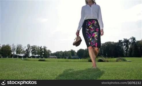 Charming businesswoman walking barefoot on green grass in the park with shoes in her hand. Attractive business lady sitting on the lawn and relaxing during break.
