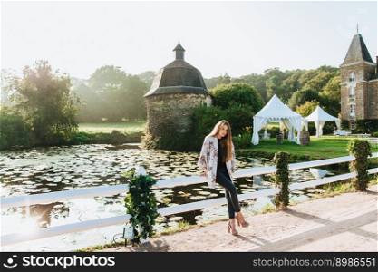 Charming calm atmosphere. Lovely slim female model wears fashionable clothes, leans at hence, poses against small lake and ancient buiduings, enjoys landscapes and landmarks, has good vacation