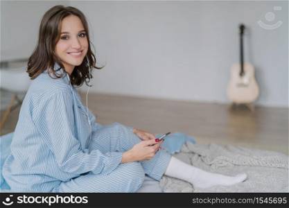Charming cheerful smiling woman musician in casual clothes, white socks, feels relaxed, listens radio or music with earphones, uses modern cell phone, poses in bed, her guitar stands in background