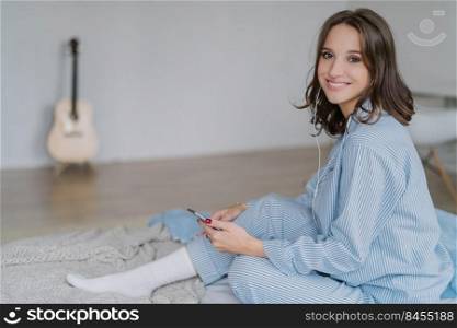 Charming cheerful smiling woman musician in casual clothes, white socks, feels relaxed, listens radio or music with earphones, uses modern cell phone, poses in bed, her guitar stands in background