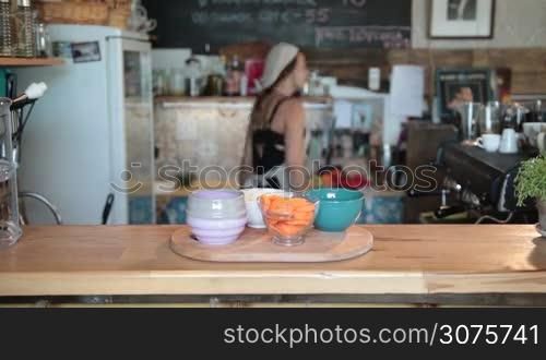 Charming female chef in white apron taking chopped ingredients in bowls on the cutting board to start cooking a soup in the kitchen