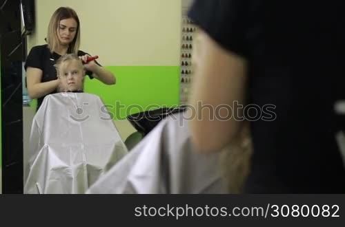 Charming female hairdresser brushing long blond girl&acute;s hair and clipping strands using barrette for fixing hairdo in barber shop. Hairstylist making new hairstyle for smiling child.