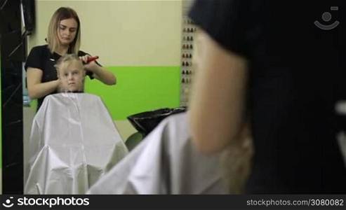 Charming female hairdresser brushing long blond girl&acute;s hair and clipping strands using barrette for fixing hairdo in barber shop. Hairstylist making new hairstyle for smiling child.