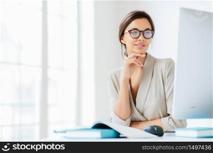 Charming female office worker keeps hand under chin, looks in screen of computer, wears spectacles for vision correction, poses in own cabinet, prepares information for future business meeting