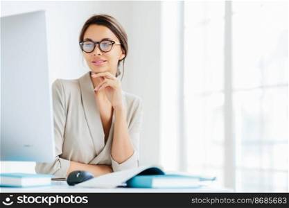 Charming female office worker keeps hand under chin, looks in screen of computer, wears spectacles for vision correction, poses in own cabinet, prepares information for future business meeting