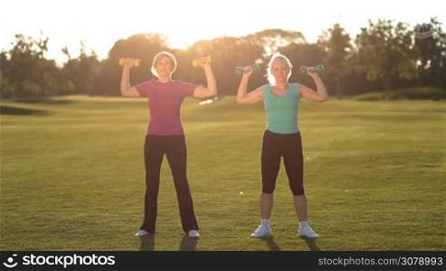 Charming fitness senior females in sports clothes lifting dumbbells while working out in the park at sunset. Smiling active sporty adult women exercising with dumbbells on green grassy field in rays of setting sun.