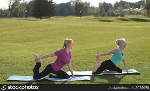 Charming fitness senior women doing pigeon asana yoga exercise on park lawn while sitting on exercise mat. Beautiful adult fit females practicing yoga, stretching in child exercise, balasana pose while working out outdoors on sunny day.
