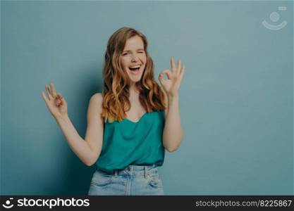 Charming funny girl showing OK sign with both hands while winking and looking at camera with big smile isolated over blue studio background. Positive body language and facial expressions concept. Charming funny girl shows OK sign with hands, winking and looking at camera with big smile