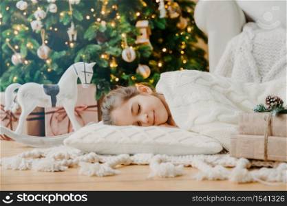 Charming girl sleeps on soft white pillow on floor against decorated New Year tree, has pleasant dreams, surrounded with toy horse and gift boxes. Children, rest and winter holidays concept.