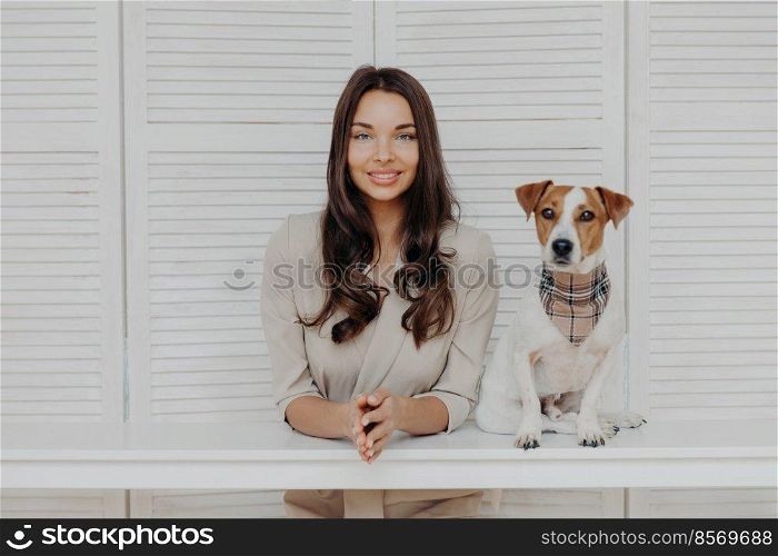 Charming gorgeous European woman with dark long hair looks directly at camera, keeps hands together, cute pedigree dog poses near, play together, have friendly relationship. Pet owner indoor