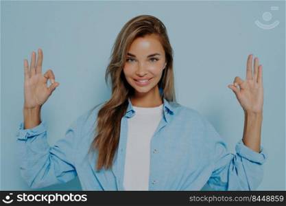 Charming happy woman in stylish casual wear making okay gesture with both hands and looking at camera with positive face expression while posing against light blue background. Charming woman making okay gesture against light blue background