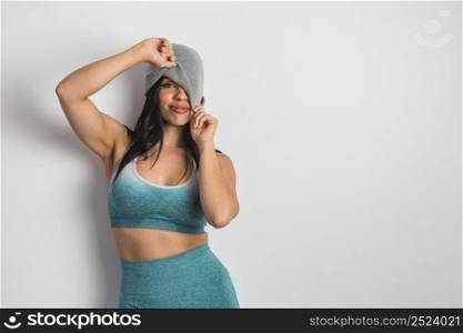 Charming Hispanic female model with long dark hair wearing sports clothes peeking out from under beanie and looking at camera on white background. Ethnic woman in sportswear pulling hat over face