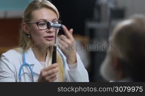 Charming long blonde female neurologist in eyesglasses testing reflexes of senior male patient &acute;s eyes wit neurological hummer during medical exam. Confident female doctor inspecting patient&acute;s nervous system using test hummer in clinic.