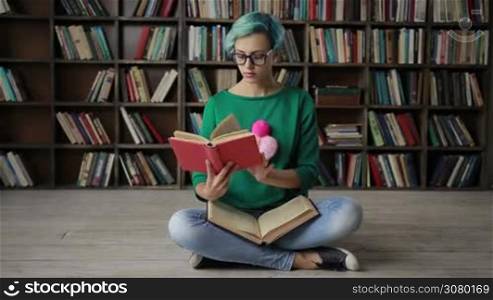 Charming nerdy woman with blue hair in spectacles sitting in lotus posture reading a book over bookshelves background. Attractive hipster girl with a book sitting cross legged, looking away thoughtfully and adjusting eyeglasses. Dolly shot. Slo mo.