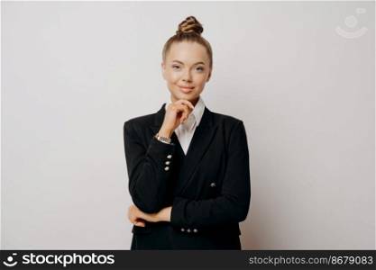 Charming professional attractive business lady in dark suit with hair in bun looking at camera with confident look, listening and thinking about opportunities, standing in front of gray background. Professional attractive business lady posing in studio