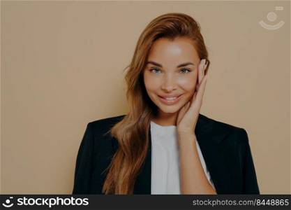 Charming prosperous young business lady dressed formally touching gently her face and smiling at camera after long working day, thinking about something pleasant while posing on beige background.. Charming business lady posing on beige background