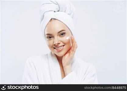 Charming relaxed beauty woman applies face cream, cares about complexion, touches cheek with hand, smiles gently at camera, dressed in bath robe, wrapped towel on washed hair, isolated on white