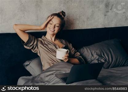 Charming relaxed young woman lying in bed at home with laptop and having morning cup of coffee, watching her favorite TV show online on computer, spending leisure time at home on weekend. Charming relaxed young woman lying in bed with laptop at home and drinking coffee