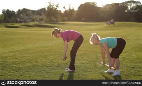 Charming sporty fit adult women performing deadlift exercise with weight body bars on park lawn at sunset. Side view. Active senior females in sport clothes doing weight lifting workout with body bars outdoors over beautiful landscape background.