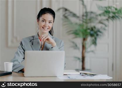Charming stylish businesswoman works freelance at laptop computer, uses high speed internet connection, holds eyeglasses in hand, enjoys favorite remote job, reads publication, sits at office desk